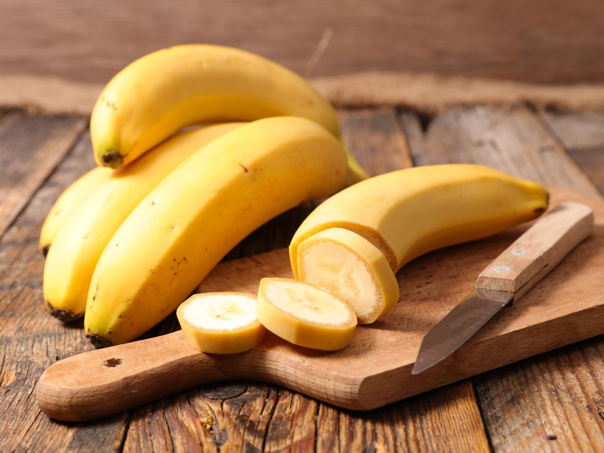 Sliced banana on a wooden cutting board with a knife next to whole bananas on a table.