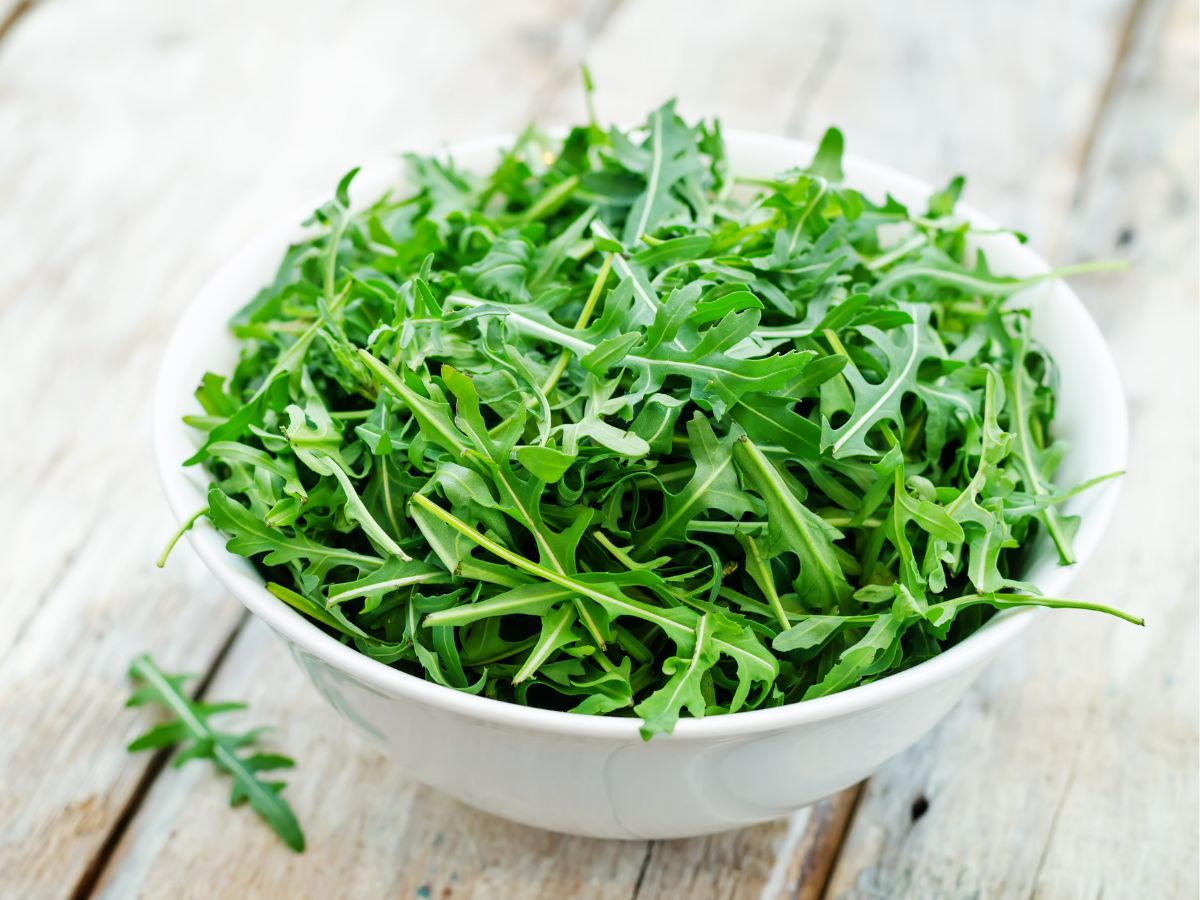 A white bowl full of arugula leaves on a wooden table.