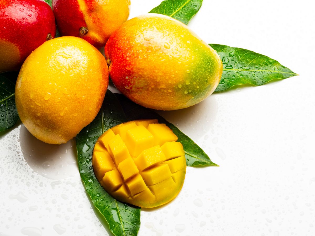 Four whole mangos and one sliced mango on mango leaves.
