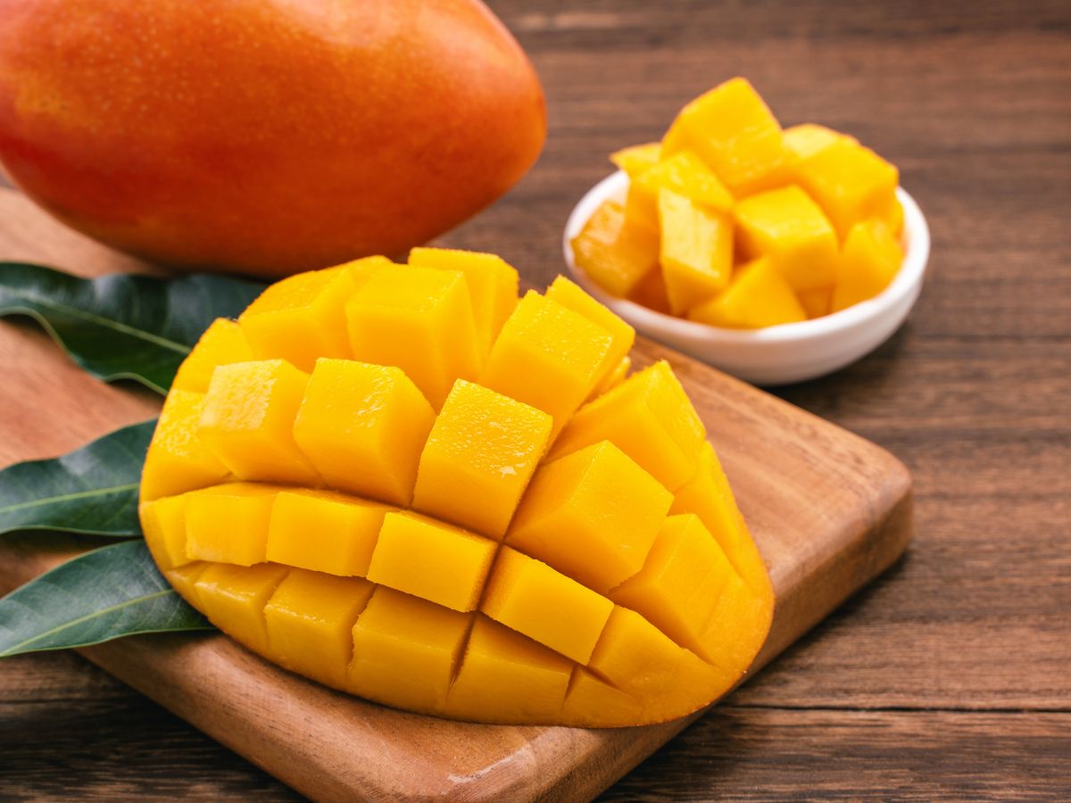 Sliced mango on a wooden cutting board and leaves next to whole mango and a bowl of sliced mango.