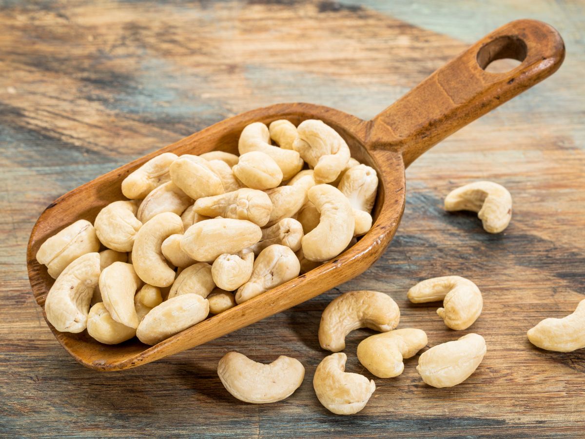 Cashew nuts in a wooden spatula on a table with scattered nuts around.