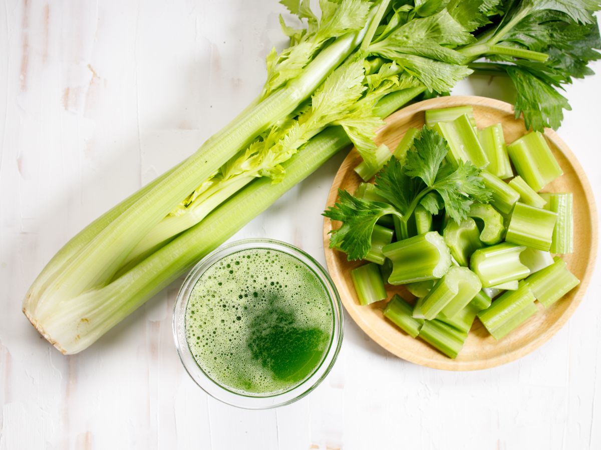 Chopped celery on a wooden plate, glass of celery juice and whole celery on a table.