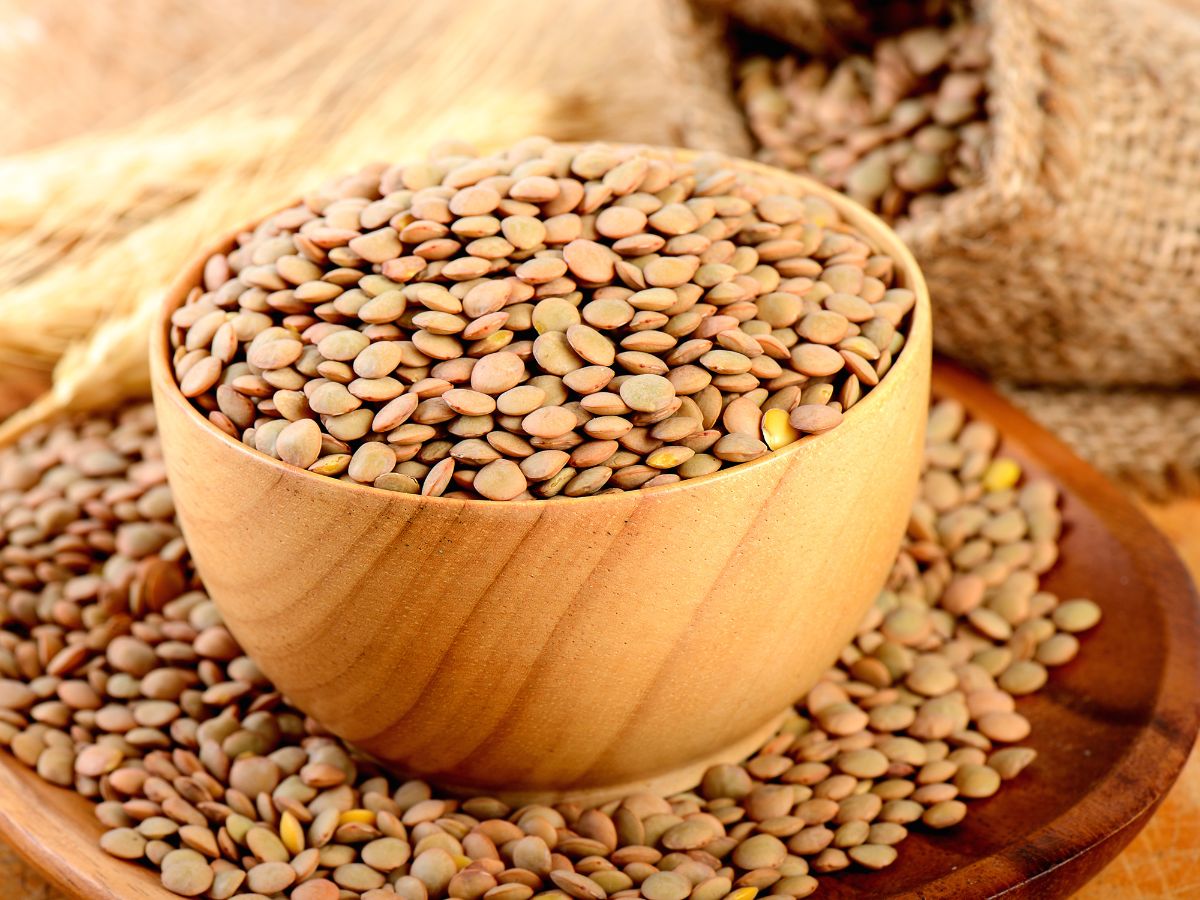 A wooden bowl full of lentils on a wooden board with scattered lentils around.