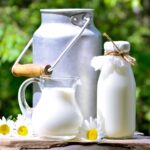 Aluminium container, glass bottle and glass cup of milk on a wooden board with white flowers.