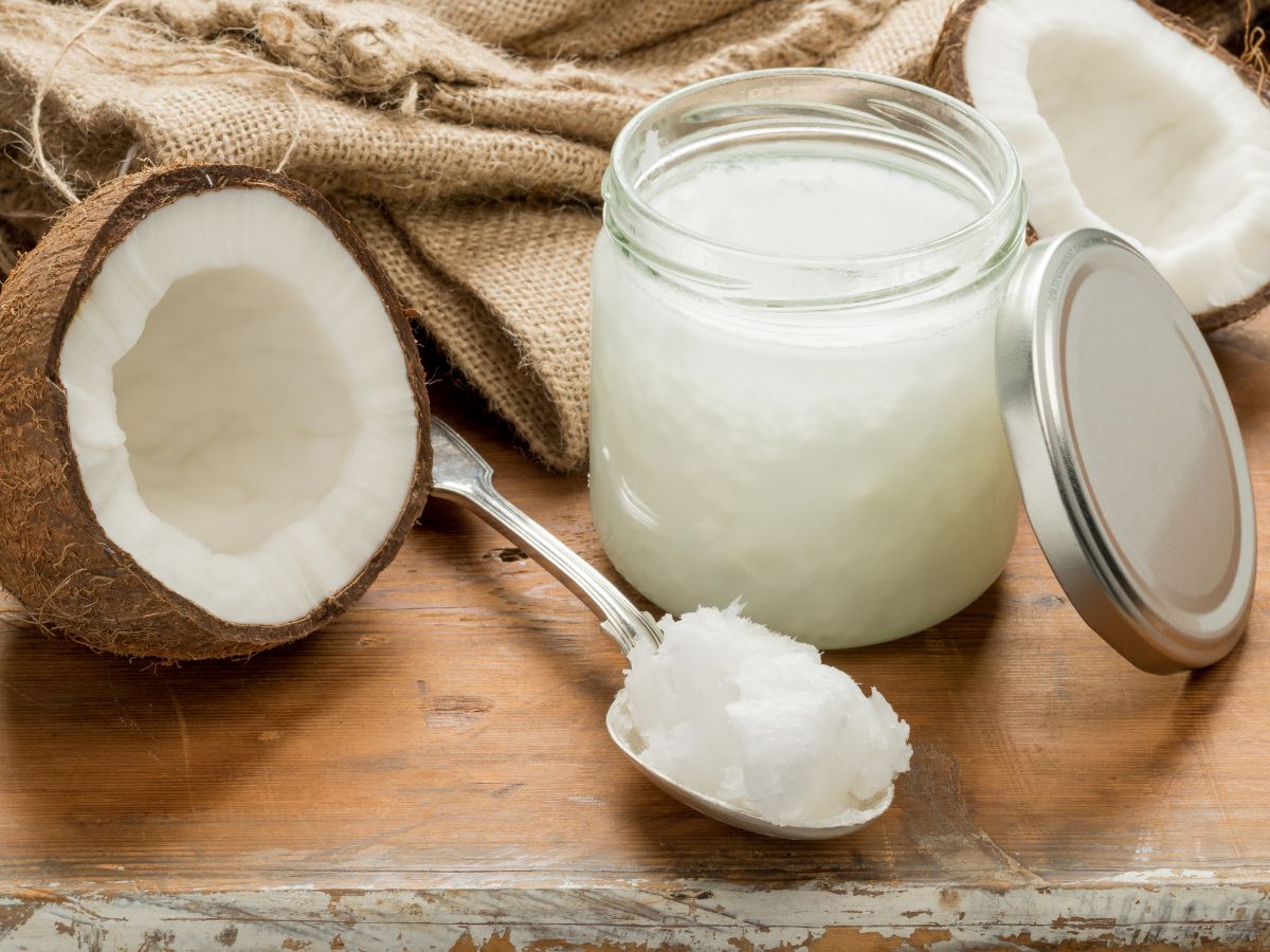 Glass jar of coconut oil on a table with coconuts and a spoon.