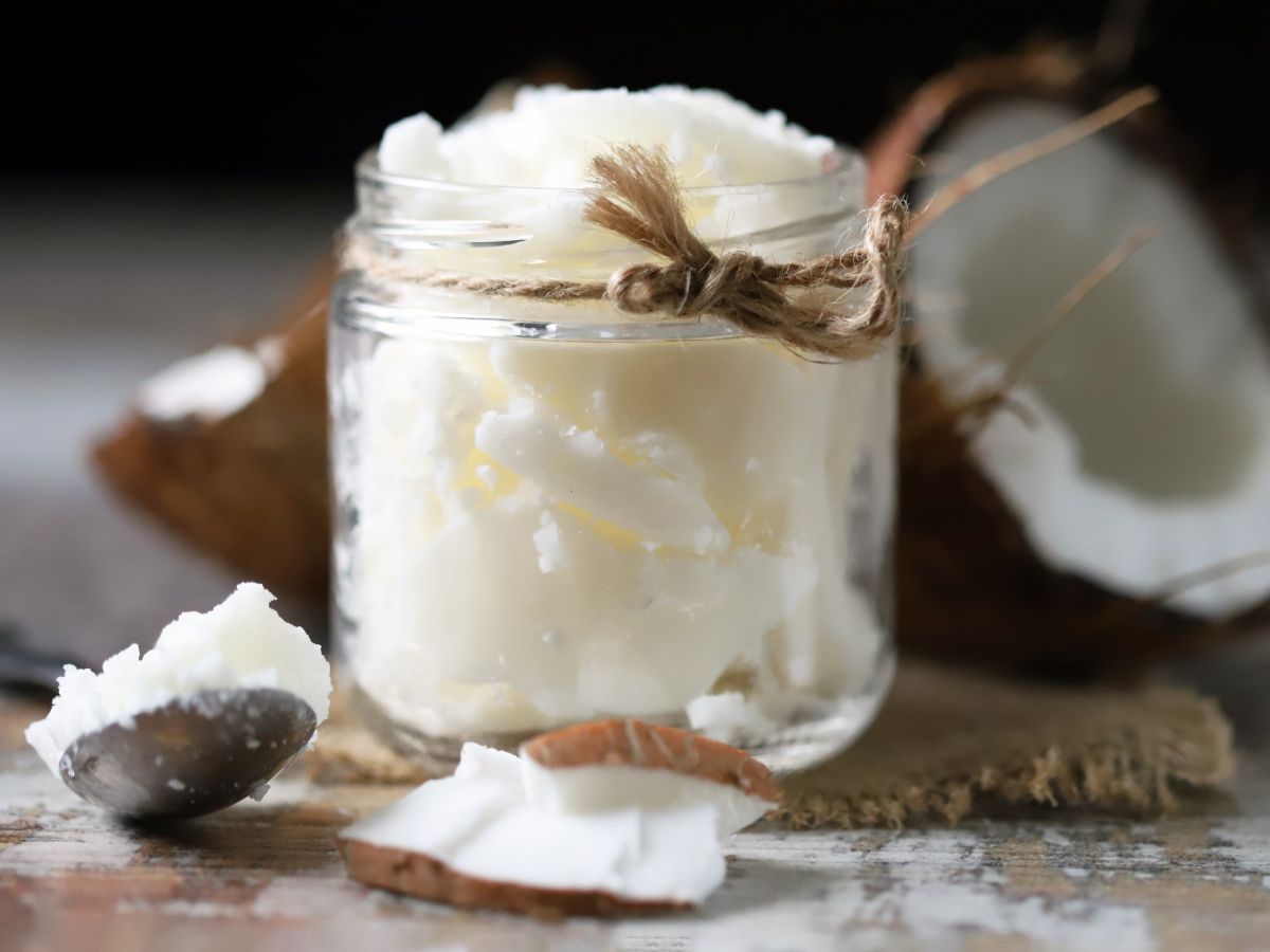 Glass jar of coconut oil on a table with a spoon and coconuts.
