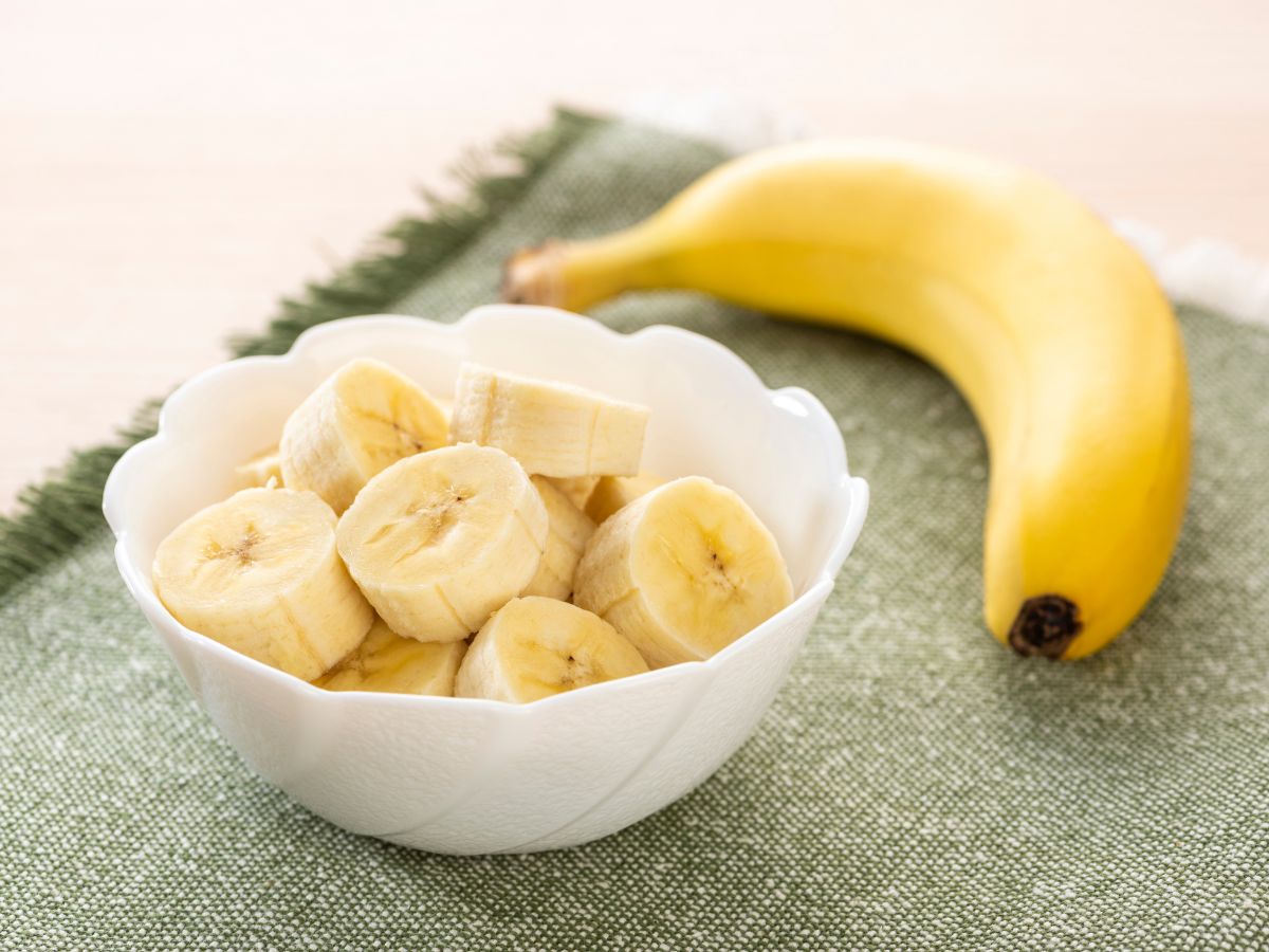 White bowl of sliced banana next to whole banana on a table.