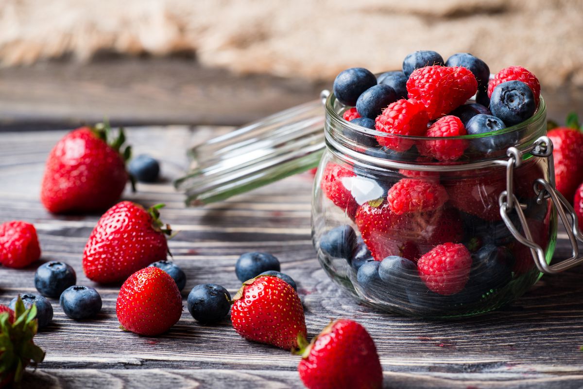 Different varieties of fresh ripe berries in a glass jar and o a wooden table.