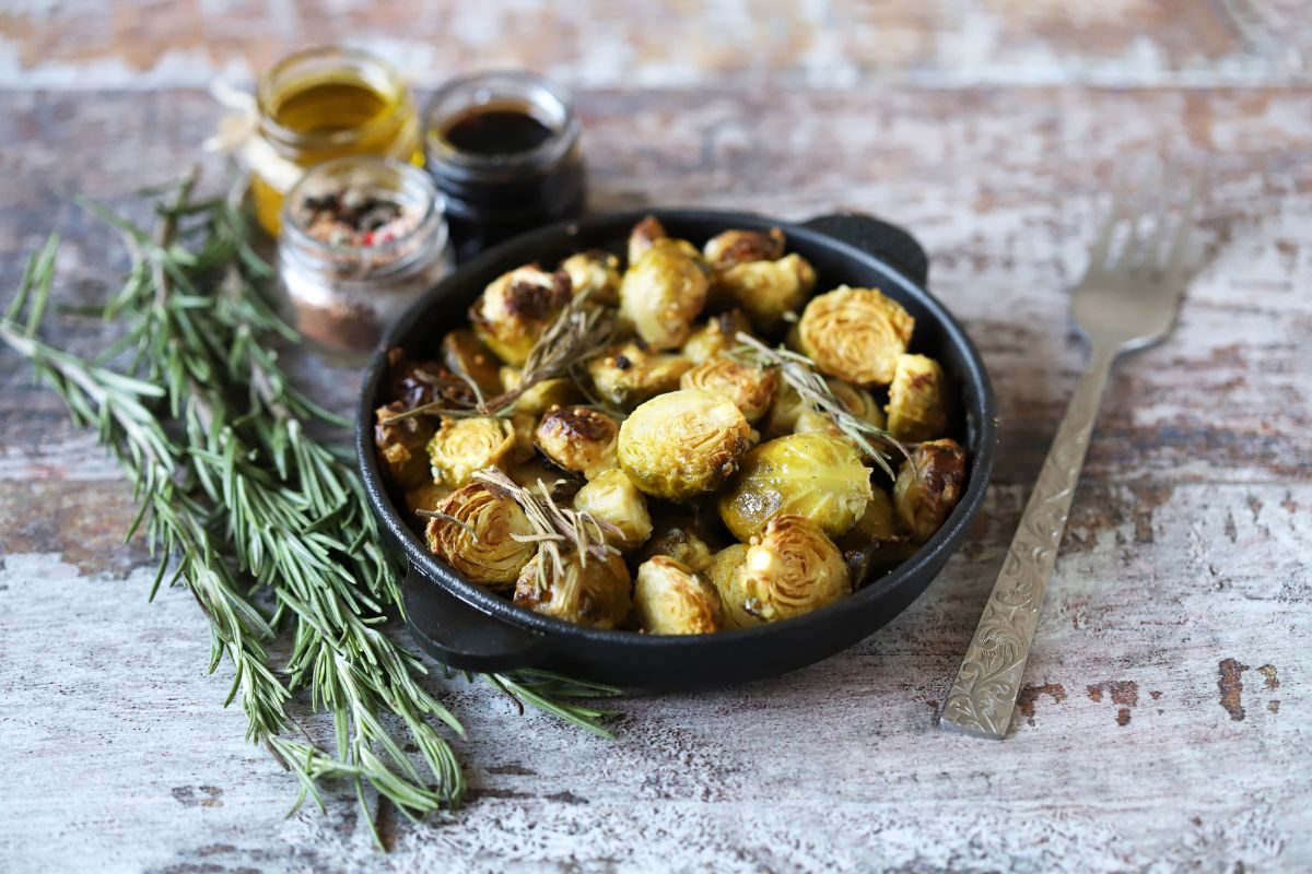 Cooked Brussel sprouts in a black bowl on a wooden table with for, condiments, and a herb.