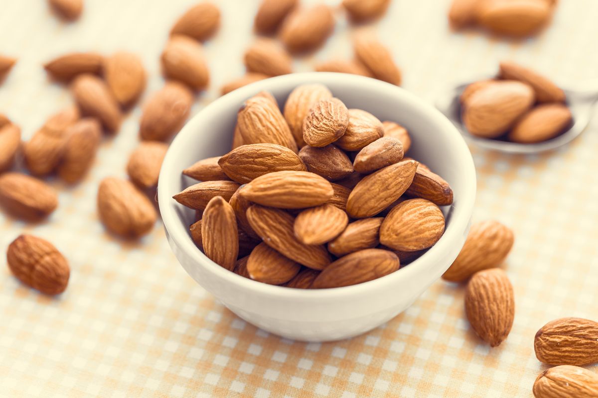 A white bowl full of almonds on a table with scattered almonds around.