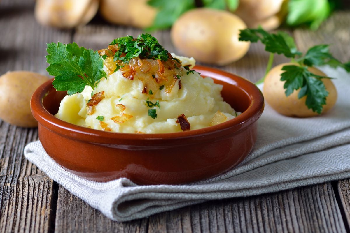 A bowl of mashed potatoes on a wooden table with scattered potatoes around.