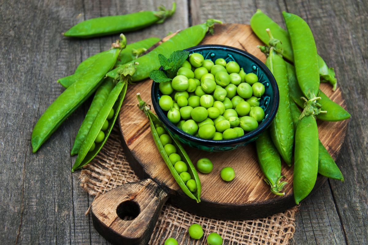 A bowl of peeled peas and whole peas on a wooden cutting board on a wooden table.
