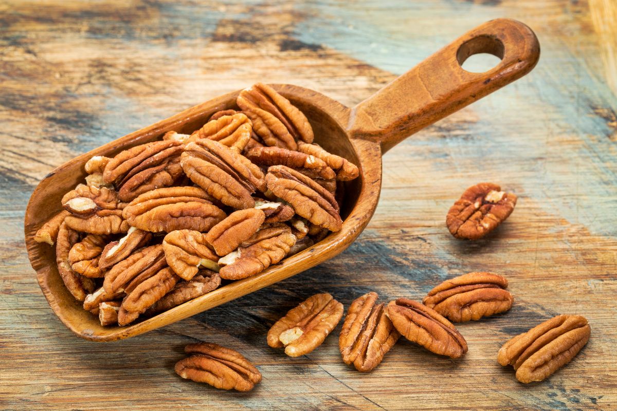 A wooden spoon full of pecans on a wooden table with scattered pecans.