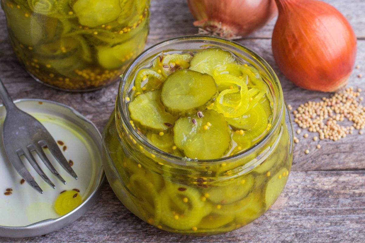A glass jar of sliced pickles on a table with a fork and onions.
