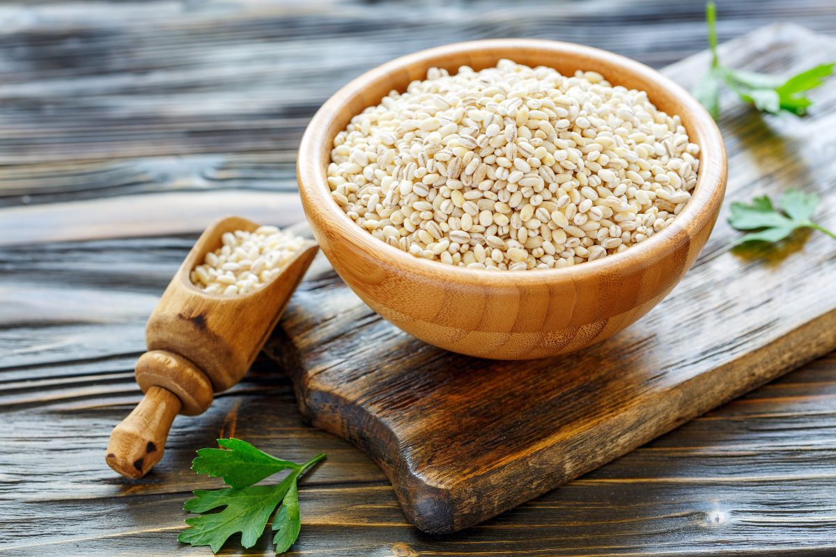 A wooden bowl full of barley on a wooden cutting board with a wooden spoon full of barley on a wooden table.
