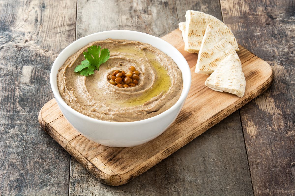 A white bowl full of hummus with pieces of bread on a wooden cutting board.