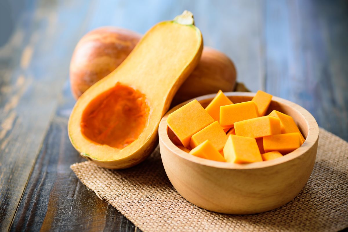 A wooden bowl of chopped butternut squash on a table with whole and half of butternut squash.