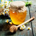 A glass jar of honey on a wooden table with flowers and a wooden dipper.