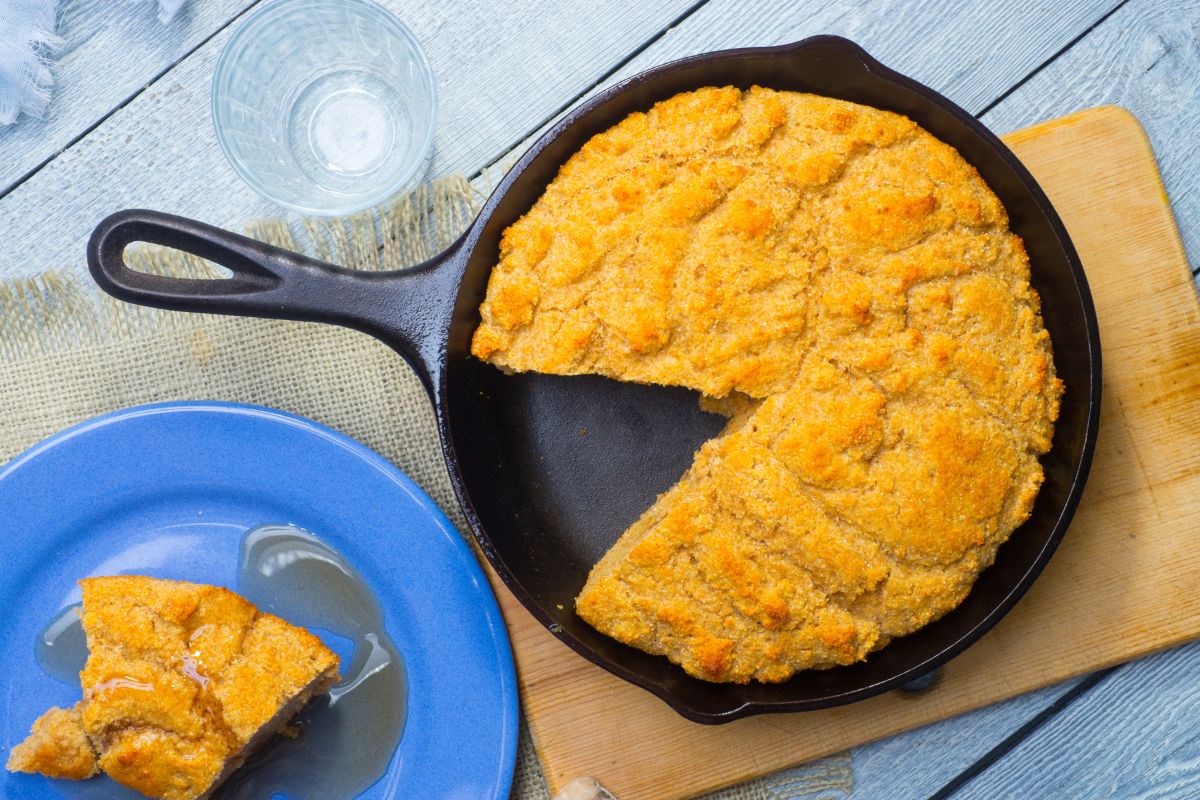 A  freshly baked cornbread on a black pan with a piece of cornbread on a blue plate.
