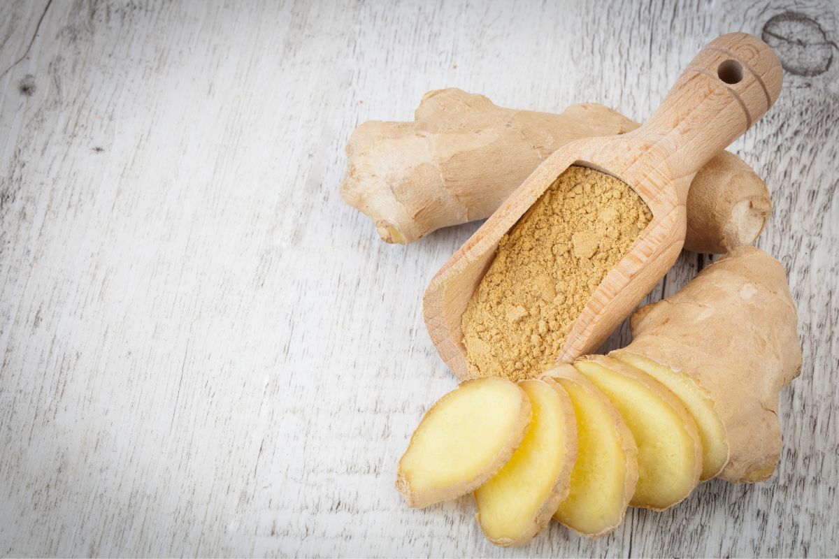 A wooden scoop of ginger and whole and sliced ginger on a table.