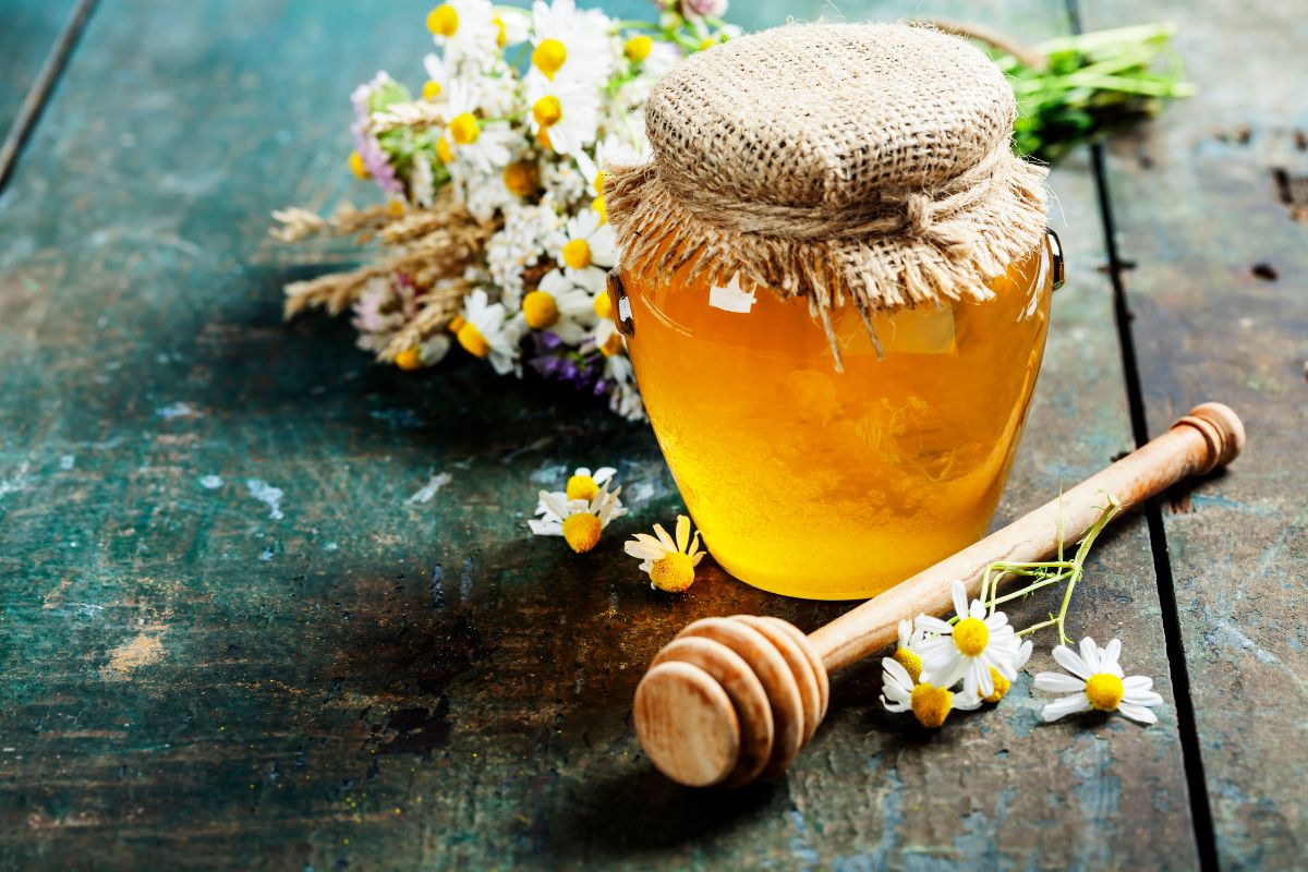 A glass jar of honey on a wooden able with flowers and a wooden dipper.