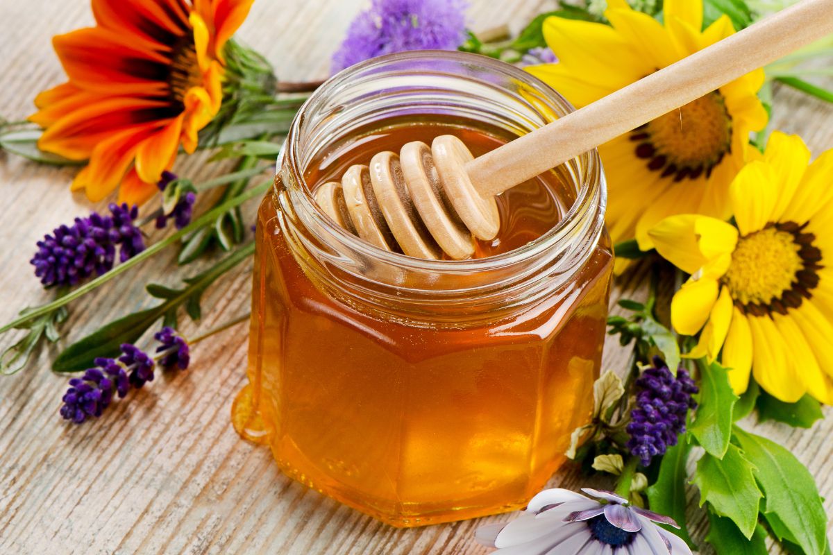 A glass jar of honey with a wooden dipper on a table with blooming flowers.