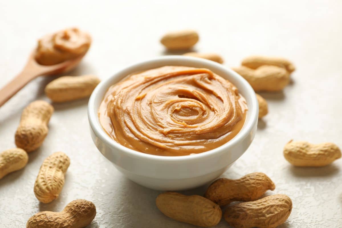 A white bowl full of peanut butter with a wooden spoon and scattered peanuts on a table.