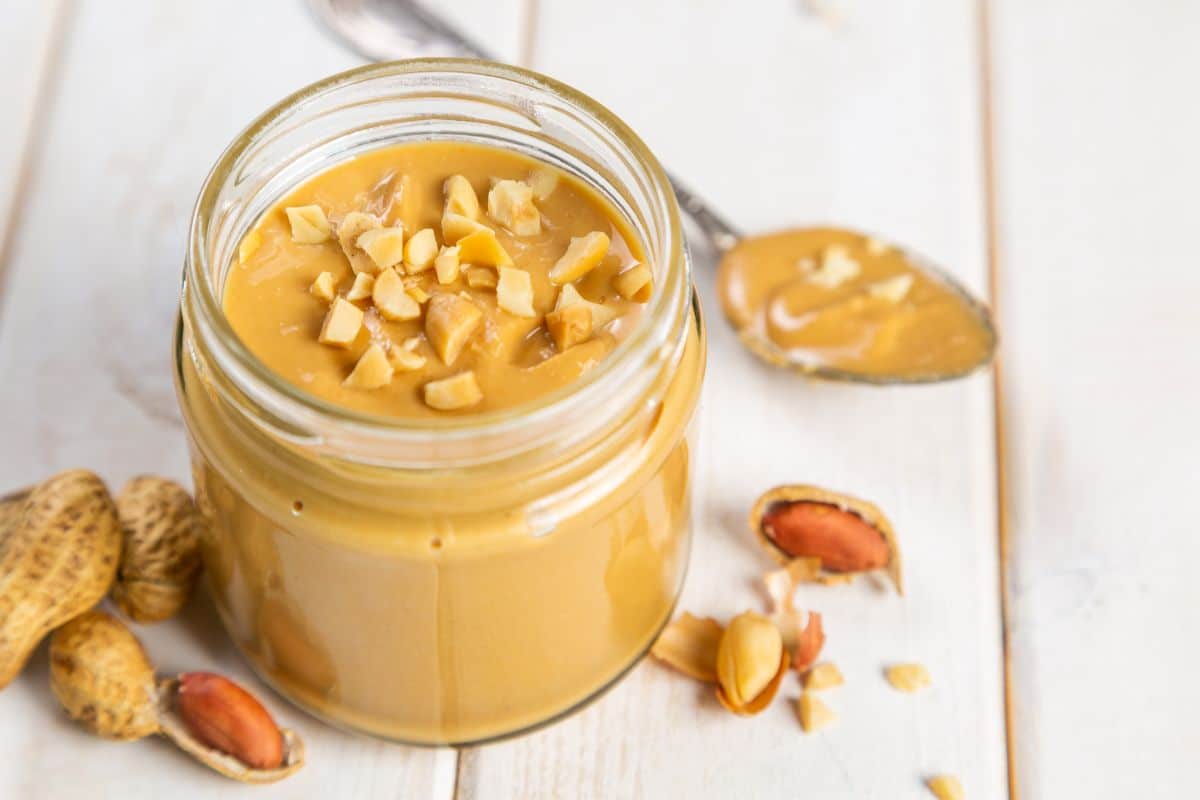 A glass jar of peanut butter with a spoon and peanuts on a wooden table.