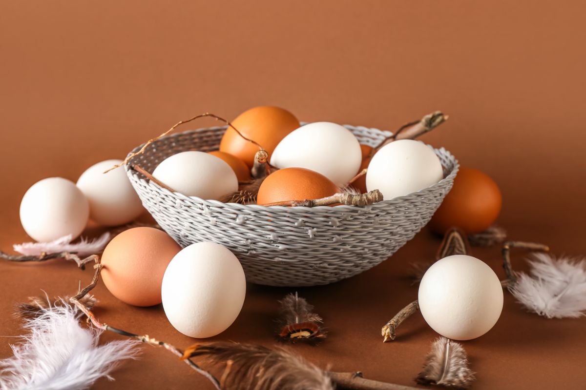 A bowl full of colorful eggs with eggs and feathers spilled on the table.