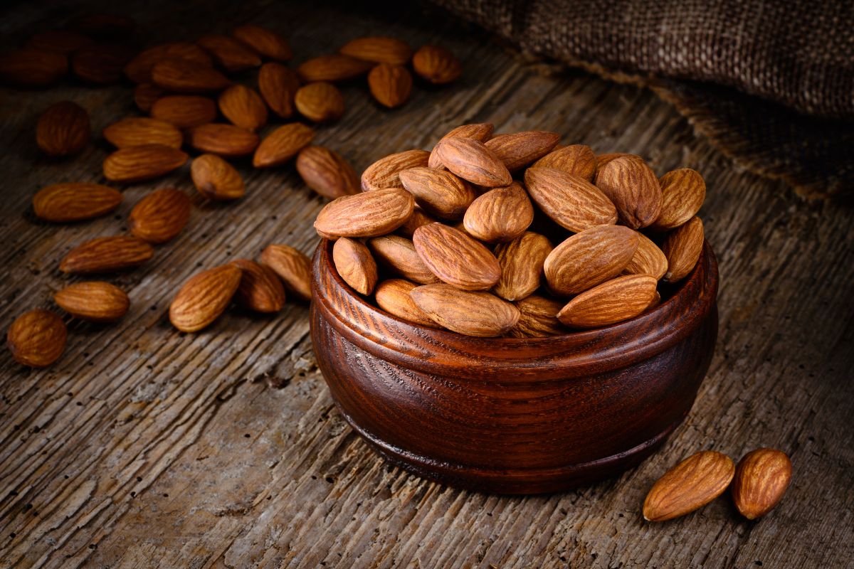 A brown bowl full of almonds on a wooden table wit scattered almonds around.