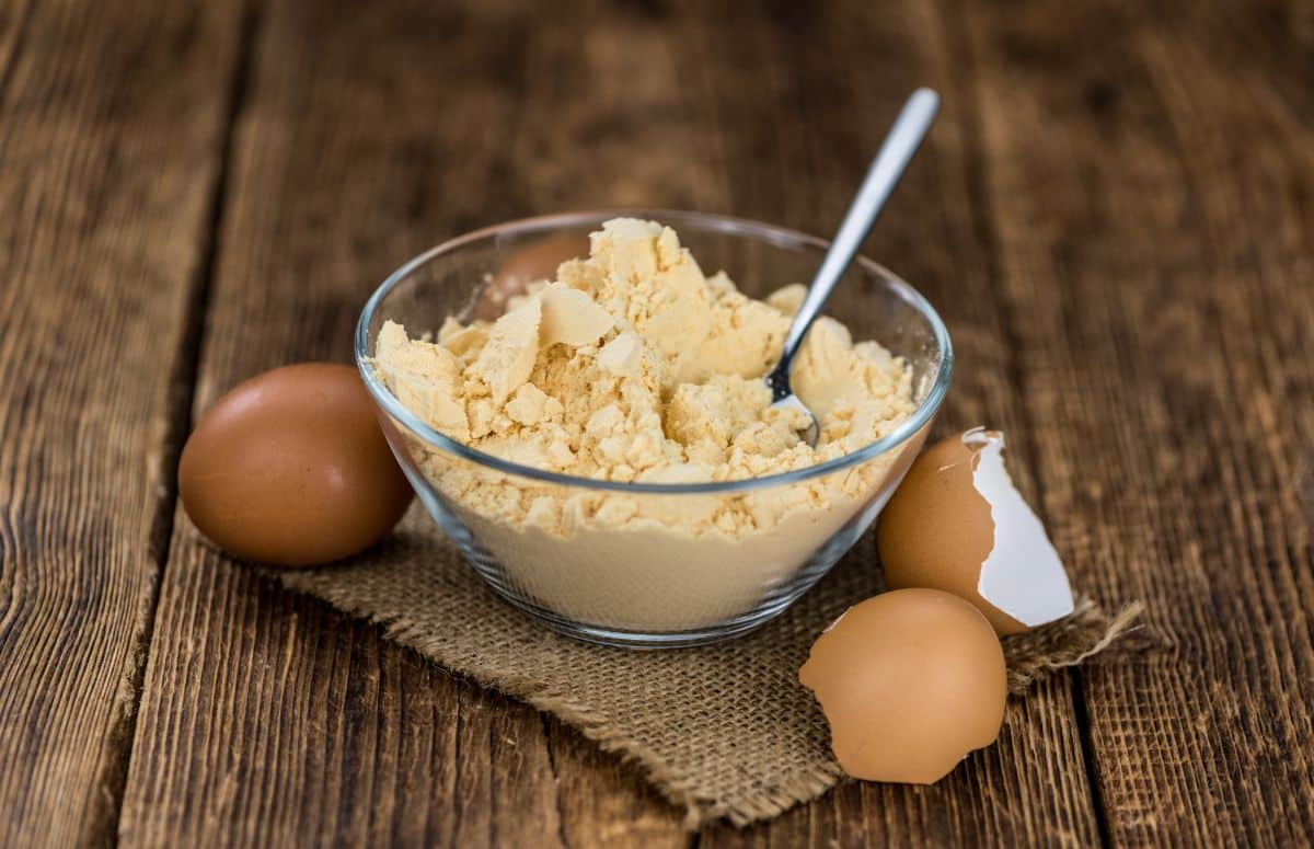 A glass bowl full of dehydrated chicken eggs with a spoon on a wooden table with egg shells.